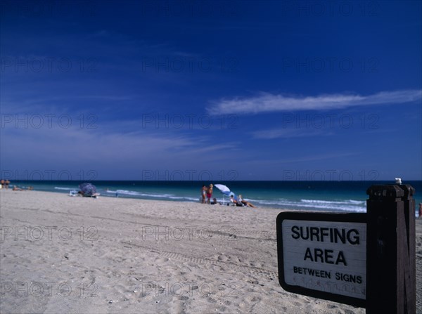 USA, Florida, Fort Lauderdale Beach, Designated Surfing Area Sign on sandy beach with sunbathers near the waters edge