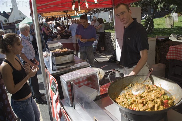 ENGLAND, West Sussex, Shoreham-by-Sea, French Market. Stallholder cooking chicken and vegetable dishes in large woks with a customer waiting