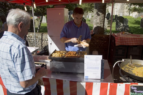 ENGLAND, West Sussex, Shoreham-by-Sea, French Market. Food stall with stallholder cooking sausage and onion baguettes with a male customer waiting