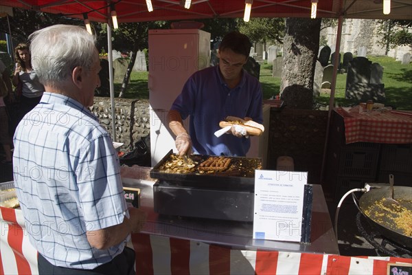 ENGLAND, West Sussex, Shoreham-by-Sea, French Market. Food stall with stallholder cooking sausage and onion baguettes with a male customer waiting