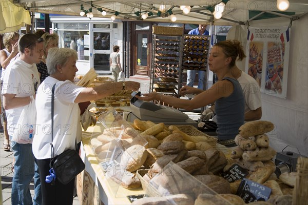 ENGLAND, West Sussex, Shoreham-by-Sea, French Market. Selection of breads on stall with customers purchasing from the stallholder