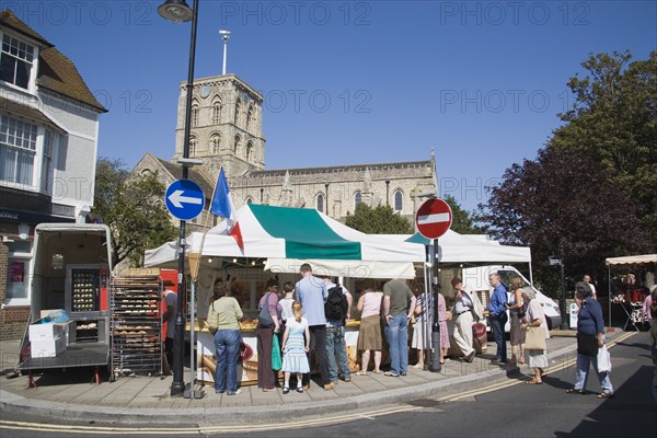 ENGLAND, West Sussex, Shoreham-by-Sea, French Market. People gathered around covered market stalls with the Church of St Mary de Haura behind.