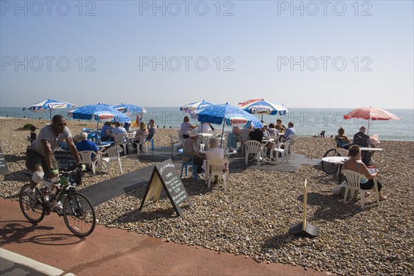 ENGLAND, West Sussex, Worthing, Beach front Cafe with people sitting at tables under parasols on the shingle overlooking the sea with a man travelling past on a bicycle