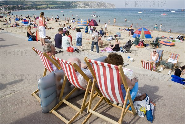 ENGLAND, Dorset, Swanage Bay, People sitting on red and white striped deckchairs looking out towards busy sandy beach with sunbathers on the sand