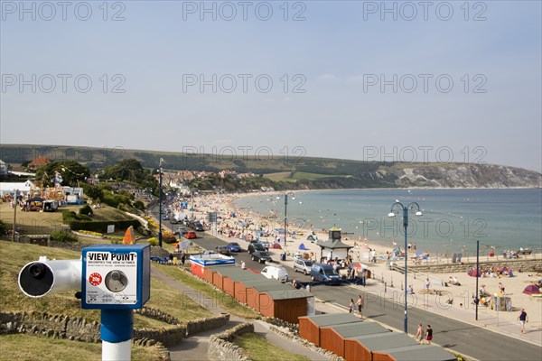 ENGLAND, Dorset, Swanage Bay, Elevated view across beach huts towards busy sandy bay with sunbathers. Viewing telescope in the foreground.