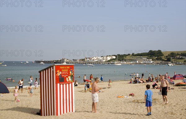 ENGLAND, Dorset, Swanage Bay, Small red and white striped Punch and Judy theatre on sandy beach with sunbathers on the sand and swimming in the sea.