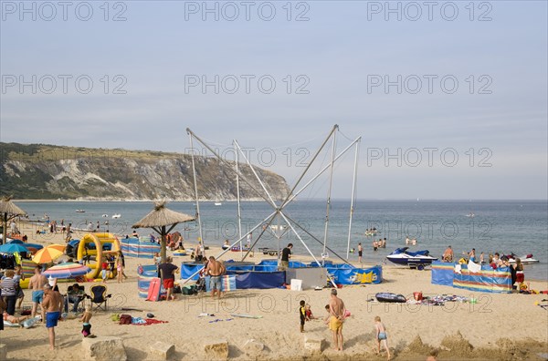ENGLAND, Dorset, Swanage Bay, Busy colourful sandy beach scattered with inflatables and windbreaks and a bungee amusement ride next to a tiki hut in the middle