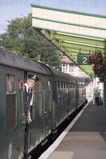 ENGLAND, Dorset, Swanage, Steam Railway Station. View along platform with train departing from station and train conductor seen looking out from carriages