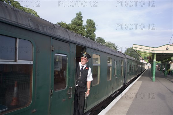 ENGLAND, Dorset, Swanage, Steam Railway Station. View along platform with train departing from station and train conductor seen looking out from carriages
