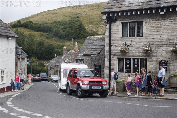 ENGLAND, Dorset, Corfe, People walking along pavements next to traditional cottages by a road with passing traffic and caravans