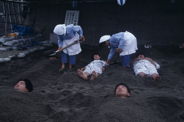 JAPAN, Kyushu, Ibusuki, Sunabulo. Hot Sand Bath with people being covered in sand by two women leaving only their head showing above the surface