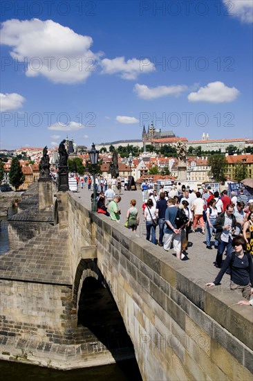 CZECH REPUBLIC, Bohemia, Prague, Tourists on the Charles Bridge across the Vltava River with Prague Castle and St Vitus' Cathedral on the horizon and light clouds in the sky