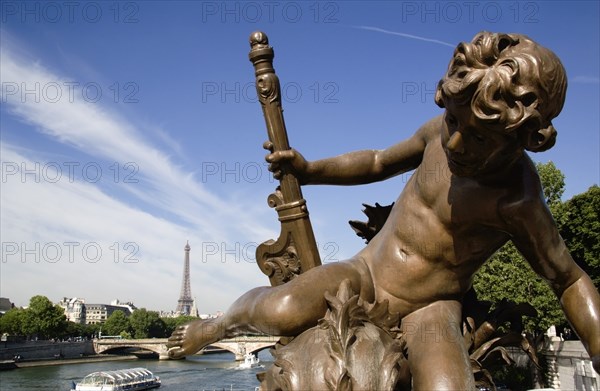 FRANCE, Ile de France, Paris, Bronze statue of a cherub on the Pont Alexandre III bridge over the River Seine with a Bateaux mouches pleasure boat with tourists passing below and the Eiffel Tower in the distance