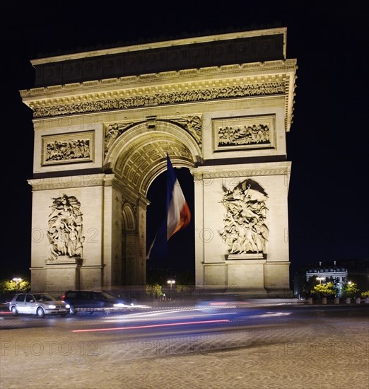 FRANCE, Ile de France, Paris, The Arc de Triomphe illuminated at night with the French tricolour flkag flying from the central arch and traffic cirlcling the Arc in the Place de Charles de Gaulle
