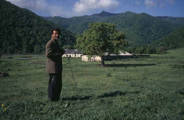 ARMENIA, Getashen, "Man standing in rural landsape with low, stone building behind."