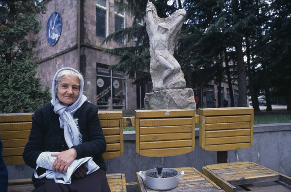 ARMENIA, Idjeran, Elderly woman selling sunflower seeds from wooden seating infront of carved stone statue.