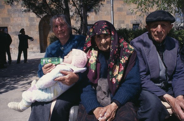 ARMENIA, Echmiadzin, Woman bottle feeding baby beside elderly couple sitting in sunshine.