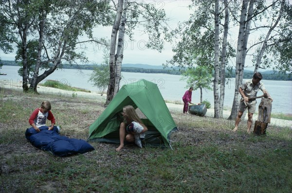CANADA, North, Camping, "Canadian family on outback holiday setting up camp beside lake.  Children with tent and sleeping bag, mother with canoe and father using axe to split log."