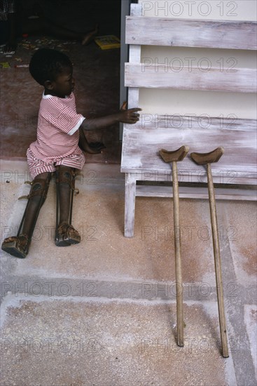 TANZANIA, Medical, Child with both legs in braces sitting in open doorway of polio unit beside pair of crutches leaning against outside wall.