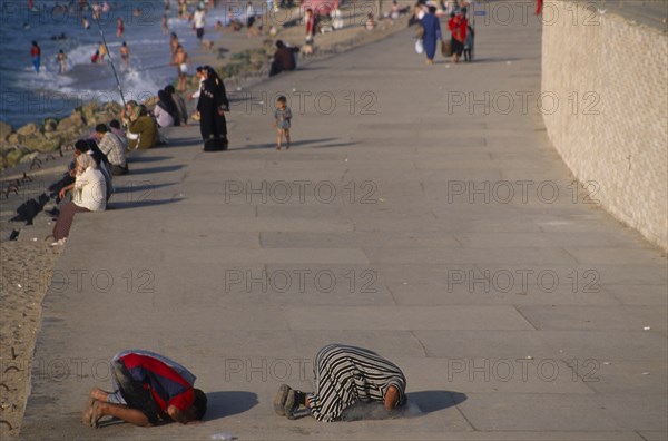 EGYPT, Nile Delta, Alexandria, Corniche Waterfront . People sitting on the edge of the promenade facing the sea with two men knelling on the ground taking part in evening prayers in the foreground.