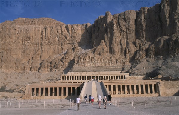 EGYPT, Nile Valley, Thebes, Deir el-Bahri. Hatshepsut Mortuary Temple. Visitors walking towards ramped entrance with limestone cliffs behind.