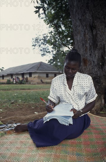 UGANDA, Iganga, Young female teacher marking exam papers outside.