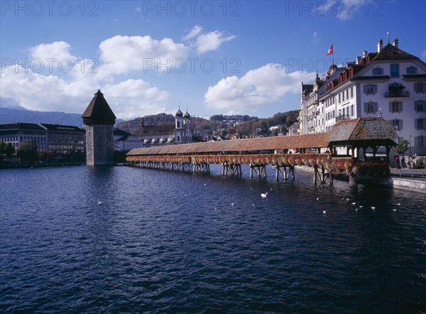 SWITZERLAND, Lucerne Central, Lucerne, Kappelbrucke flower decked covered bridge spanning across The River Reuss. Adjoining the bridge is the Wasserturm 13th Century octagonal tower