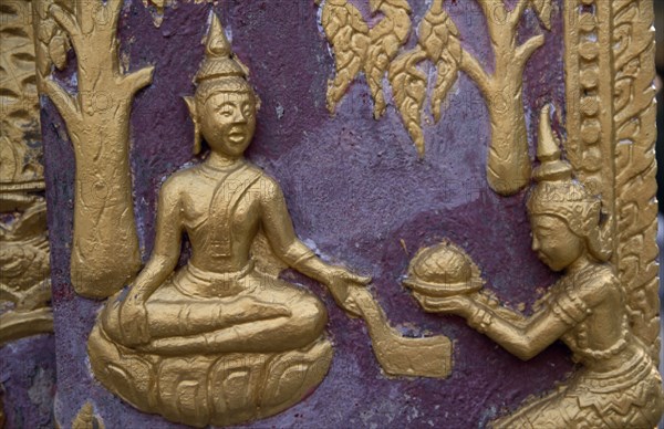 LAOS, Luang Prabang, Wat Xieng Thong.  Temple detail of gold painted relief carving on purple background depicting seated Buddha receiving offerings.
