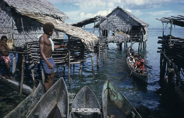 PACIFIC ISLANDS, Melanesia, Solomon Islands, Thatched stilt houses on artificial island with man and child on wooden bridge and approaching canoe.  Part view of wooden canoes in immediate foreground.
