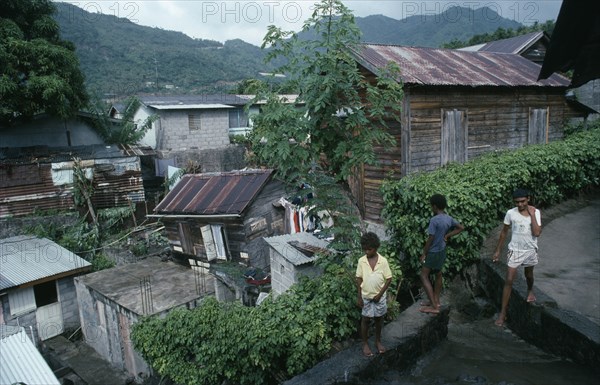 ST LUCIA, Soufriere, "Wood and breeze block housing with corrugated tin rooves, hanging washing and children standing on  drainage channel in foreground."