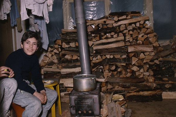 ARMENIA, Yerevan, Boy sitting beside wood burning stove in house with large stack of wood behind during winter time.