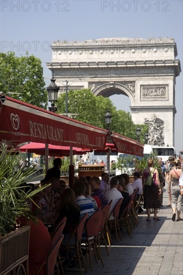 FRANCE, Ile de France, Paris, Tourists eating lunch at tables under shade in the Champs Elysees with the Arc de Triomphe in the distance