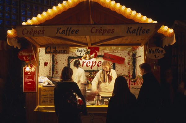 GERMANY, Berlin, "Breitscheidplatz. Christmas Market. Customers waiting at food stall illuminated at night cooking and selling Crepes, Coffee and Gluhwein which is similar to mulled wine."