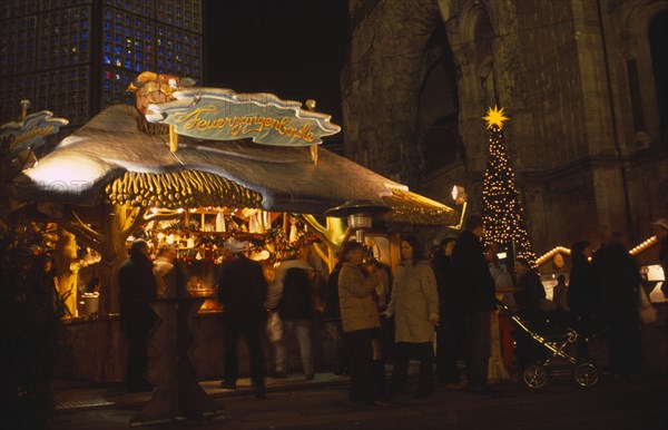 GERMANY, Berlin, "Breitscheidplatz. Christmas Market. People gathered around an Illuminated stall selling Feuerzangenbowle a German alcoholic drink made with wine, rum and sugar loaf.  A Christmas tree with a star nearby."