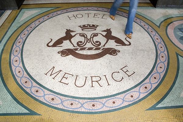 FRANCE, Ile de France, Paris, Woman walking across a mosaic with a crest and the words Hotel Meurice on the pavement outside the entrance door to the five star hotel in the Rue de Rivoli