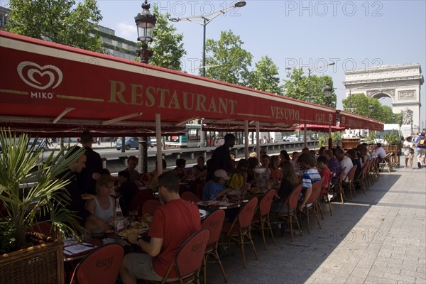 FRANCE, Ile de France, Paris, Tourists eating lunch at tables under shade in the Champs Elysees with the Arc de Triomphe in the distance