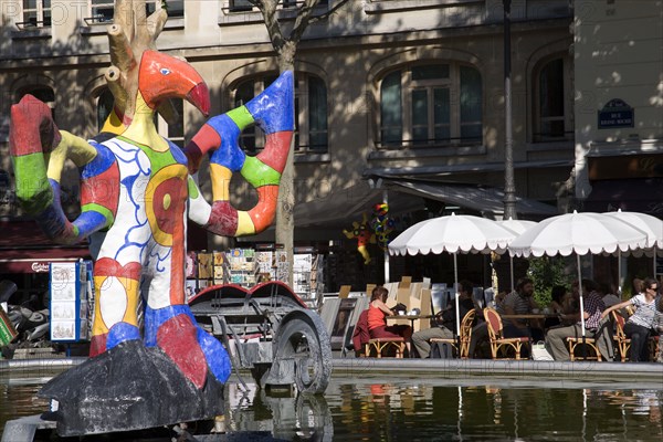 FRANCE, Ile de France, Paris, People sitting at tables under umbrellas beside the colourful contemporary fountains by Niki de Saint Phalle and Jean Tinguely in Place Igor Stravinsky beside the Pompidou Centre in Beauborg Les Halles