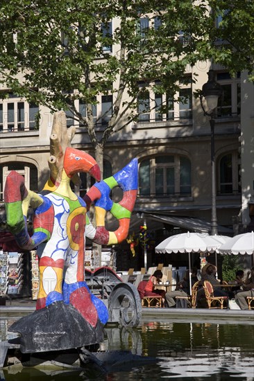 FRANCE, Ile de France, Paris, People sitting at tables under umbrellas beside the colourful contemporary fountains by Niki de Saint Phalle and Jean Tinguely in Place Igor Stravinsky beside the Pompidou Centre in Beauborg Les Halles