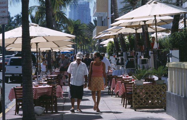 USA, Florida, Miami, South Beach. Ocean Drive. A man and woman walking past a sidewalk cafe restaurant with table and chairs under sun umbrellas