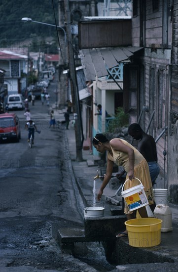 ST LUCIA, Soufriere, Man and woman collecting water from standpipe in narrow street with open gutter.