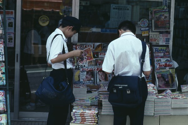 JAPAN, Chiba, Narita, Two Junior High Japanese schoolboys in uniform reading magazines from pavement stand with typical ‘cute girl’ covers.