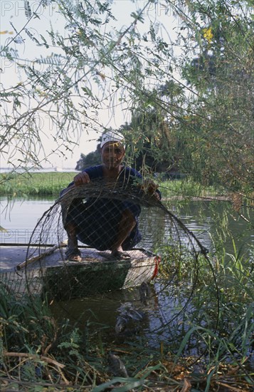 EGYPT, Luxor, Nile Fisherman crouching down next to river bank on the West Bank under tree branches displaying his catch of fish