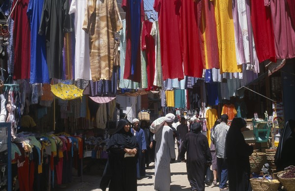 EGYPT, Nile Valley, Luxor, Sharia el-Souk street in the main market / bazaar. Men and women walking amongst market stalls under hanging displays of brightly coloured clothing