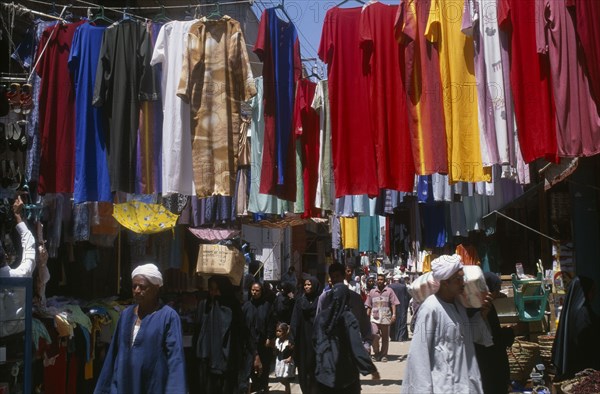 EGYPT, Nile Valley, Luxor, Sharia el-Souk street in the main market / bazaar. Men and women walking amongst market stalls under hanging displays of brightly coloured clothing