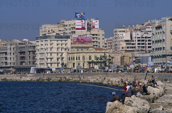 EGYPT, Nile Delta, Alexandria, Corniche Waterfront . Men and boys fishing from rocks over the sea next to the promenade. Overlooked by tall buildings behind