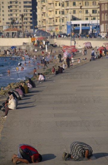EGYPT, Nile Delta, Alexandria, Corniche Waterfront . People sitting on the edge of the promenade facing the sea with two men knelling on the ground taking part in evening prayers in the foreground.
