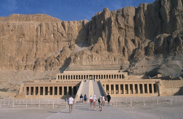 EGYPT, Nile Valley, Thebes, Deir el-Bahri. Hatshepsut Mortuary Temple. Visitors walking towards ramped entrance with limestone cliffs behind