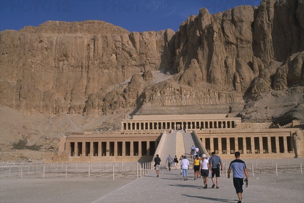 EGYPT, Nile Valley, Thebes, Deir el-Bahri. Hatshepsut Mortuary Temple. Visitors walking towards ramped entrance with limestone cliffs behind