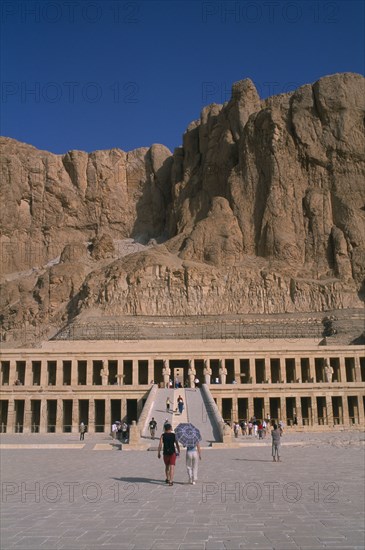 EGYPT, Nile Valley, Thebes, Deir el-Bahri. Hatshepsut Mortuary Temple. Visitors walking towards ramped entrance with limestone cliffs behind
