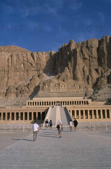 EGYPT, Nile Valley, Thebes, Deir el-Bahri. Hatshepsut Mortuary Temple. Visitors walking towards ramped entrance with limestone cliffs behind.